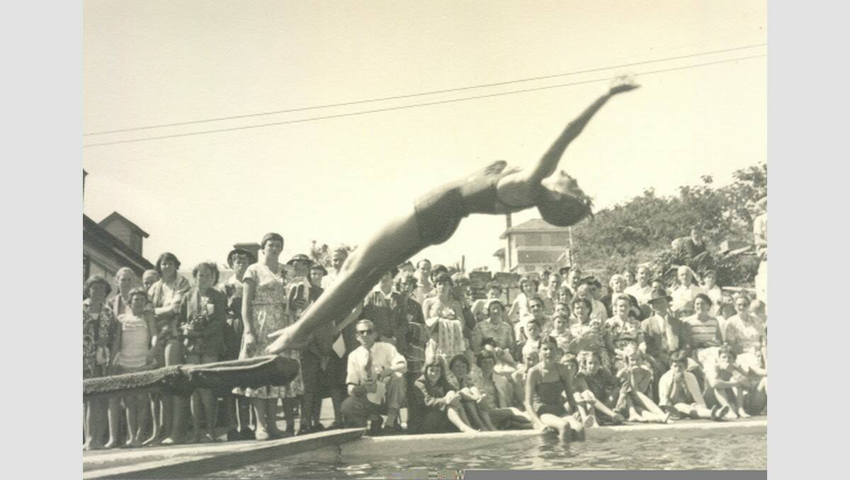 A crowd watches a dive attempt at the Technical School Swimming Sports in 1956 at the swimming baths in Gilles Street. The oval baths edge is still visible at the site but the area is grassed over. Mozart Hall is the building on the left of the photo. SOURCE: Warrnambool & District Historical Society.